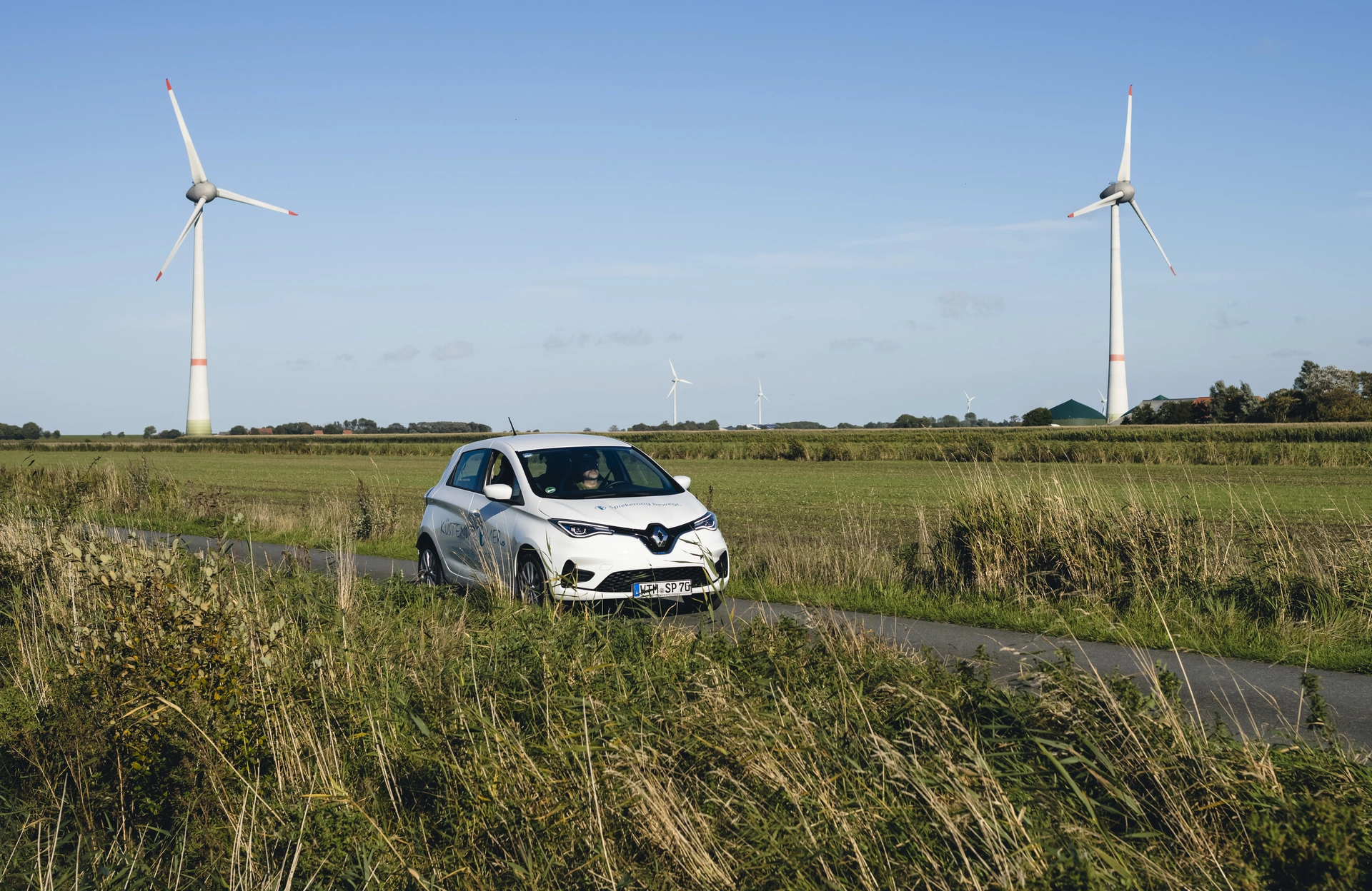 Ein Auto fährt über eine Landstraße, im Hintergrund sind zwei Windräder zu sehen.